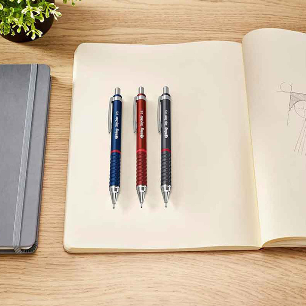 Three Rotring Tikky Retro 0.7mm Mechanical Pencil in 3 colours on an open notebook with a wooden surface and plant in the background